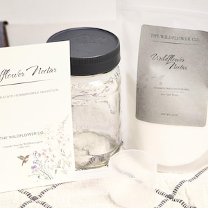 Puede incluir: Un tarro de cristal transparente con tapa negra, un paquete de mezcla de néctar para colibríes, una cuchara medidora y una tarjeta con el texto "Wildflower Nectar". El paquete y la tarjeta tienen el texto "The Wildflower Co."