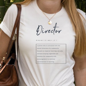 May include: White t-shirt with the word "Director" printed in blue script. The definition of the word is printed below in black text: "a person who is entrusted with the overall direction of a community focused on classical learning, one who oversees keeping organized and blankets the community with encouragement to continue conversations in learning."