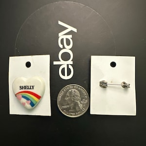 May include: A white ceramic heart-shaped pin with a rainbow and the word "SHELLY" in pink lettering. The pin is attached to a silver safety pin.