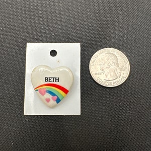 May include: White ceramic heart-shaped pin with the name "Beth" written in red. The heart has a rainbow and three pink hearts on it. The pin is displayed on a white card with a hole at the top.