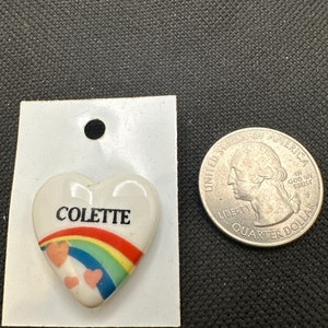 May include: White ceramic heart-shaped pin with a rainbow and two small hearts. The pin says "Colette" in black lettering.