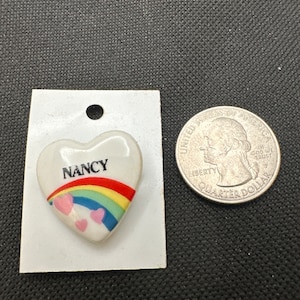 May include: White heart-shaped ceramic pin with a rainbow and three pink hearts. The pin says "NANCY" in black letters.