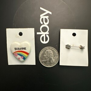 May include: A white porcelain heart-shaped pin with a rainbow and the word "SUSANNE" on it. The pin is on a white card with a hole at the top. A silver safety pin is on a separate white card.