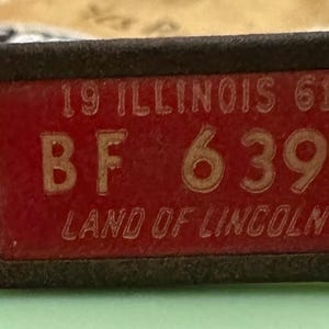 May include: A red metal keychain with a black border and a white inscription that reads "19 Illinois 61 BF 6 399 Land of Lincoln".