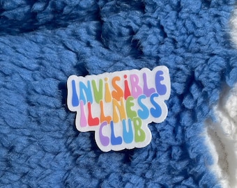 Op de afbeelding: Een sticker met de tekst "Invisible Illness Club" in regenboogkleuren op een blauwe en witte pluche achtergrond.