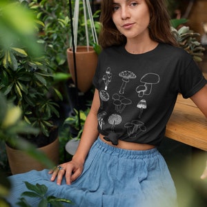 May include: A black t-shirt with a white graphic of various mushrooms. The model is wearing the shirt and a blue skirt. The model is sitting on a wooden bench in a garden setting.