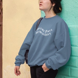 A young woman leaning against a light green wall with a steel blue teal crewneck sweatshirt on that has wavy text that says "This is my flare day sweatshirt". She is looking away and looks kind of tired.