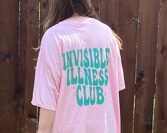Può includere: Una maglietta rosa chiaro oversize con la scritta "INVISIBLE ILLNESS CLUB" in verde. La maglietta è realizzata in materiale morbido e viene indossata all'aperto. Lo sfondo comprende una staccionata in legno ed erba verde.