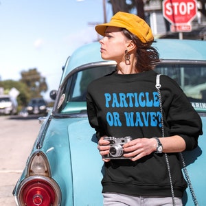 May include: Black sweatshirt with the text "PARTICLE OR WAVE?" in blue, worn by a person. The person is wearing a yellow hat and holding a vintage camera. A classic turquoise car is in the background.