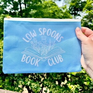 May include: A light blue zippered pouch with the text "Low Spoons Book Club" in white. The text is surrounded by a white outline of a book with flowers.