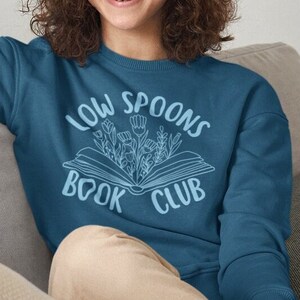 May include: A teal blue sweatshirt with the text "Low Spoons Book Club" in white lettering. The text is surrounded by a stylized image of an open book with flowers growing out of it.