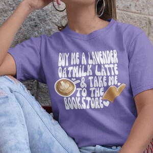 May include: A woman wearing a purple t-shirt with the text "Buy me a lavender oatmilk latte & take me to the bookstore" in a retro font. The shirt features a graphic of a coffee cup and an open book.