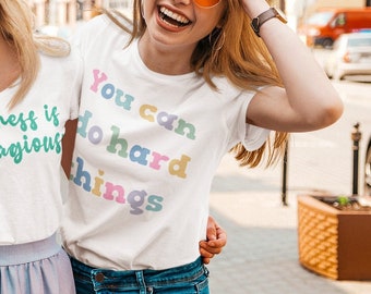 May include: Two women wearing white t-shirts with colorful text. The woman on the left's shirt says "kindness is contagious" and the woman on the right's shirt says "You can do hard things".