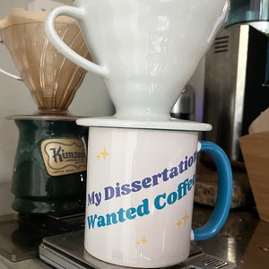 Op de afbeelding: Witte keramische koffiemok met blauwe en paarse tekst die "My Dissertation Wanted Coffee" met gele sterren zegt. De mok staat op een digitale keukenweegschaal.