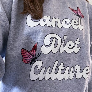 Pu&ograve; includere: Felpa grigia con il testo "Cancel Diet Culture" in lettere bianche con contorno. Due farfalle rosa sono sulla felpa.