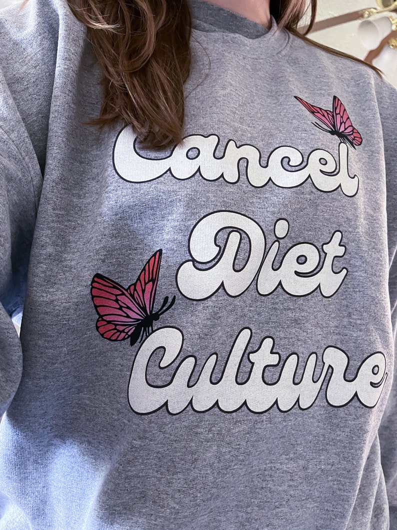 Pu&ograve; includere: Felpa grigia con il testo "Cancel Diet Culture" in lettere bianche con contorno. Due farfalle rosa sono sulla felpa.