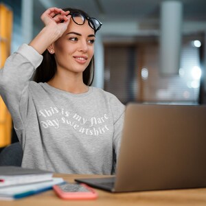 A young woman at a laptop is wearing a light ash grey crewneck sweatshirt that has wavy text that says "This is my flare day sweatshirt". She has school books in front of her and looks like she might be studying the desk.