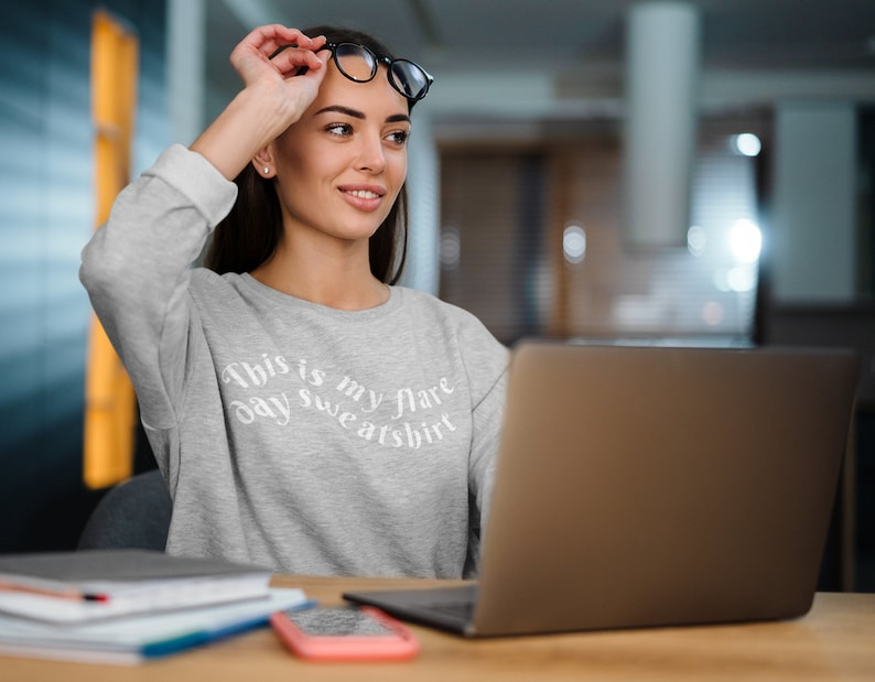 A young woman at a laptop is wearing a light ash grey crewneck sweatshirt that has wavy text that says "This is my flare day sweatshirt". She has school books in front of her and looks like she might be studying the desk.