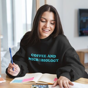 Puede incluir: Una mujer lleva una sudadera negra con el texto "Coffee and Microbiology" en letras azules. Está sentada en una mesa con un cuaderno y un bolígrafo.