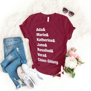 May include: A maroon t-shirt with white text that reads "Ada & Marie & Katherine & Jane & Rosalind & Vera & Chien-Shiung".