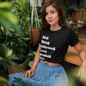 May include: A woman wearing a black cropped t-shirt with white text that reads "Ada & Marie & Katherine & Jane & Rosalind & Vera". She is wearing a blue skirt and sitting on a wooden surface.