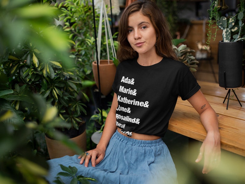 May include: A woman wearing a black cropped t-shirt with white text that reads "Ada & Marie & Katherine & Jane & Rosalind & Vera". She is wearing a blue skirt and sitting on a wooden surface.