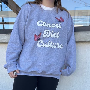Pu&ograve; includere: Felpa grigia con il testo "Cancel Diet Culture" in bianco con grafiche di farfalle rosa.