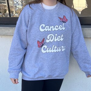 Pu&ograve; includere: Una felpa grigia con il testo "Cancel Diet Culture" in bianco con un font retr&ograve;. Due farfalle rosa sono stampate sulla felpa.