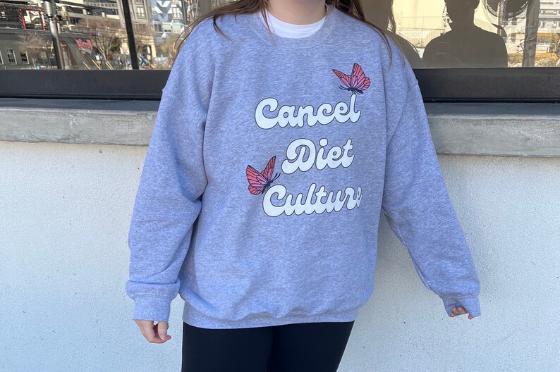 Pu&ograve; includere: Una felpa grigia con il testo "Cancel Diet Culture" in bianco con un font retr&ograve;. Due farfalle rosa sono stampate sulla felpa.