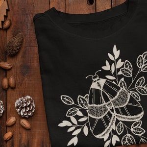 Op de afbeelding: Zwart T-shirt met een wit mot- en bloemenontwerp. De mot heeft ingewikkelde details en is omringd door bladeren.