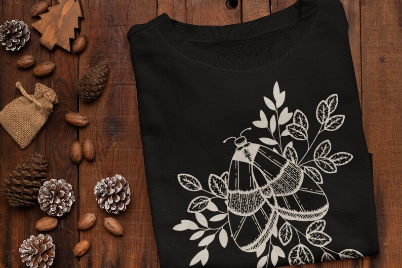 Op de afbeelding: Zwart T-shirt met een wit mot- en bloemenontwerp. De mot heeft ingewikkelde details en is omringd door bladeren.