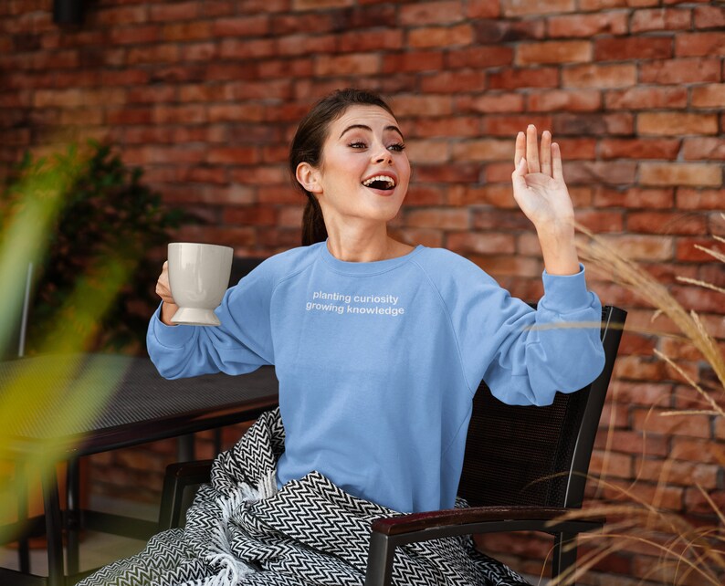 Puede incluir: Una mujer con una sudadera azul claro con el texto "planting curiosity growing knowledge" sentada en una silla con una manta. Est&aacute; sosteniendo una taza blanca en su mano izquierda y su mano derecha est&aacute; levantada.