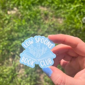 Peut inclure: Un autocollant bleu clair avec un contour blanc d'un livre ouvert avec des fleurs qui en sortent. Le texte "Low Spoons Book Club" est écrit autour du livre.