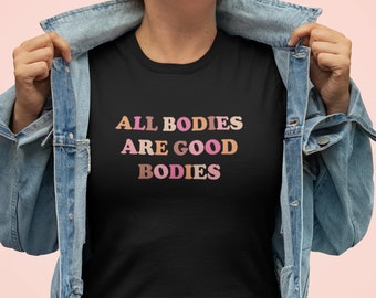 May include: A black t-shirt with the text "ALL BODIES ARE GOOD BODIES" in colorful letters. The shirt is being held by a person wearing a denim jacket.