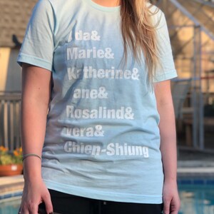 May include: A light blue t-shirt with white text that reads "da& Marie& Katherine& ane& Rosalind& vera& Chien-Shiung".