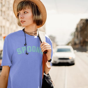 A woman with a hat on is wearing a purple oversized tee shirt in the color black that says in teal letters "SPOONIE". She is crossing the street.