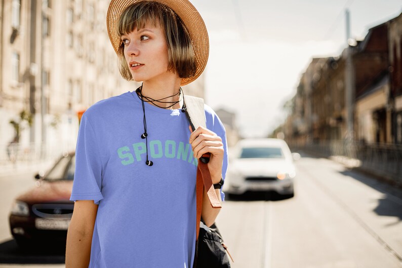 A woman with a hat on is wearing a purple oversized tee shirt in the color black that says in teal letters "SPOONIE". She is crossing the street.