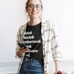 May include: A woman wearing a black t-shirt with white text that reads "Ada & Marie & Katherine & ... & ... & ..."  She is also wearing a white and black plaid shirt, blue jeans, and glasses. She is holding a black smartphone in her left hand and using a laptop computer with her right hand.
