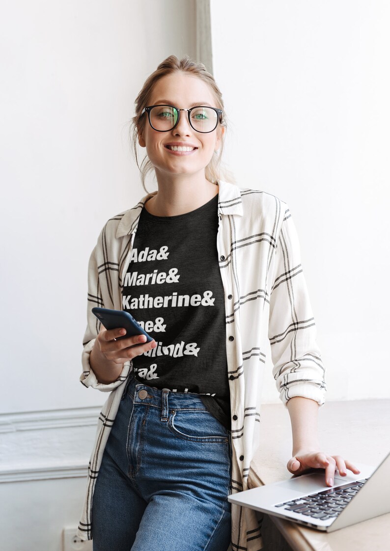 May include: A woman wearing a black t-shirt with white text that reads "Ada & Marie & Katherine & ... & ... & ..."  She is also wearing a white and black plaid shirt, blue jeans, and glasses. She is holding a black smartphone in her left hand and using a laptop computer with her right hand.