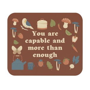May include: A brown background with the text "You are capable and more than enough" in a retro font. The text is surrounded by various illustrations of mushrooms, a bird, a teapot, a ladybug, a carrot, a tomato, a turnip, a lemon, a bell pepper, a strawberry, and leaves.