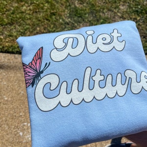 Pu&ograve; includere: Felpa blu chiaro con il testo "Diet Culture" in bianco, con caratteri stilizzati. Una farfalla rosa con segni neri &egrave; sul lato sinistro della felpa.