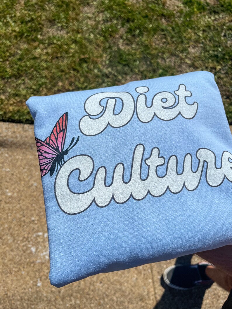 Pu&ograve; includere: Felpa blu chiaro con il testo "Diet Culture" in bianco, con caratteri stilizzati. Una farfalla rosa con segni neri &egrave; sul lato sinistro della felpa.