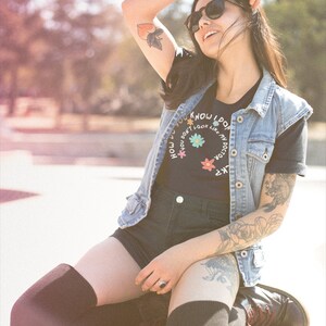 May include: A woman wearing a black t-shirt with the text "How do you know I don't look like my doctor?" in a circle with flowers, a denim vest, black shorts, and black thigh-high socks. She is sitting on a concrete surface with her arm raised and sunglasses on.