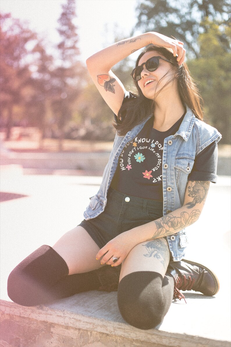 May include: A woman wearing a black t-shirt with the text "How do you know I don't look like my doctor?" in a circle with flowers, a denim vest, black shorts, and black thigh-high socks. She is sitting on a concrete surface with her arm raised and sunglasses on.