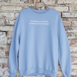 Puede incluir: Una sudadera de color azul claro con el texto "planting curiosity growing knowledge" impreso en blanco en la parte delantera.