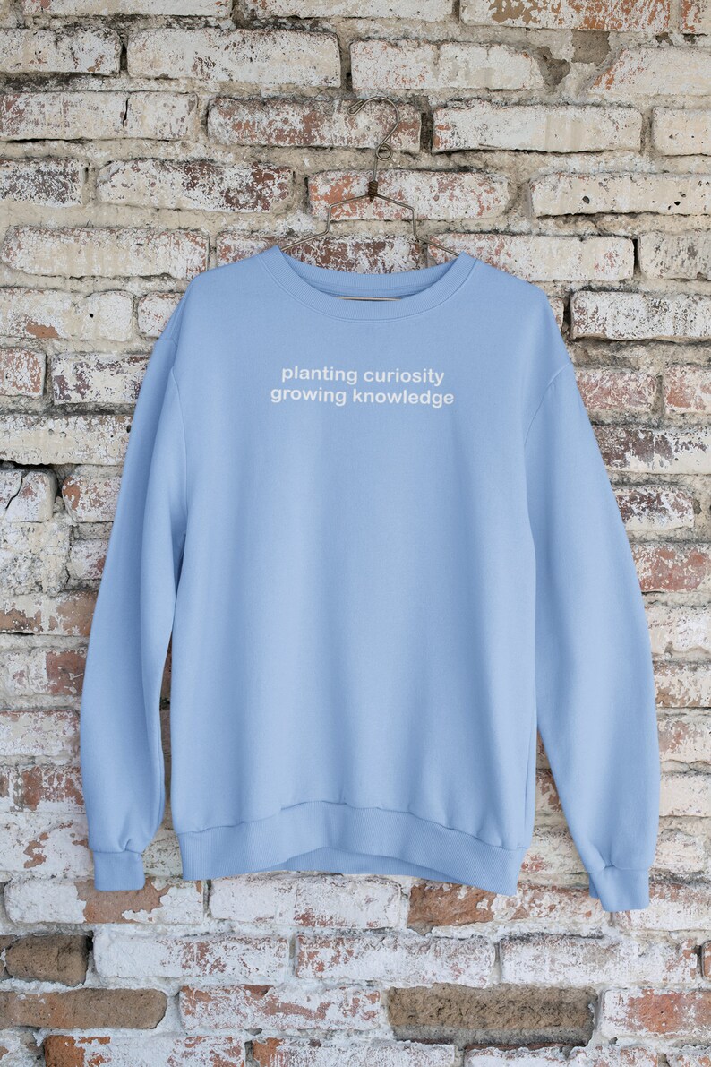 Puede incluir: Una sudadera de color azul claro con el texto "planting curiosity growing knowledge" impreso en blanco en la parte delantera.