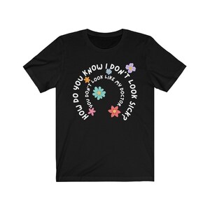 May include: Black t-shirt with a white circle design featuring the text "How do you know I don't look sick?" with colorful flowers around the text.