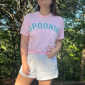 May include: A pink t-shirt with the word "SPOONIE" printed in teal green letters. The shirt is worn by a person standing in a forest.