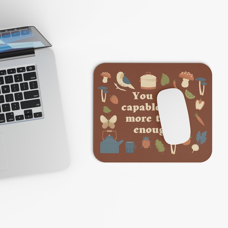 May include: A brown mouse pad with a colorful illustration of a butterfly, teapot, mushrooms, and other nature-themed elements. The text "You are capable of more than enough" is written in a retro font.