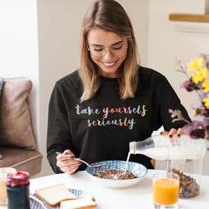 May include: A woman wearing a black sweatshirt with the text "take yourself seriously" in rainbow colors pours milk into a bowl of cereal.
